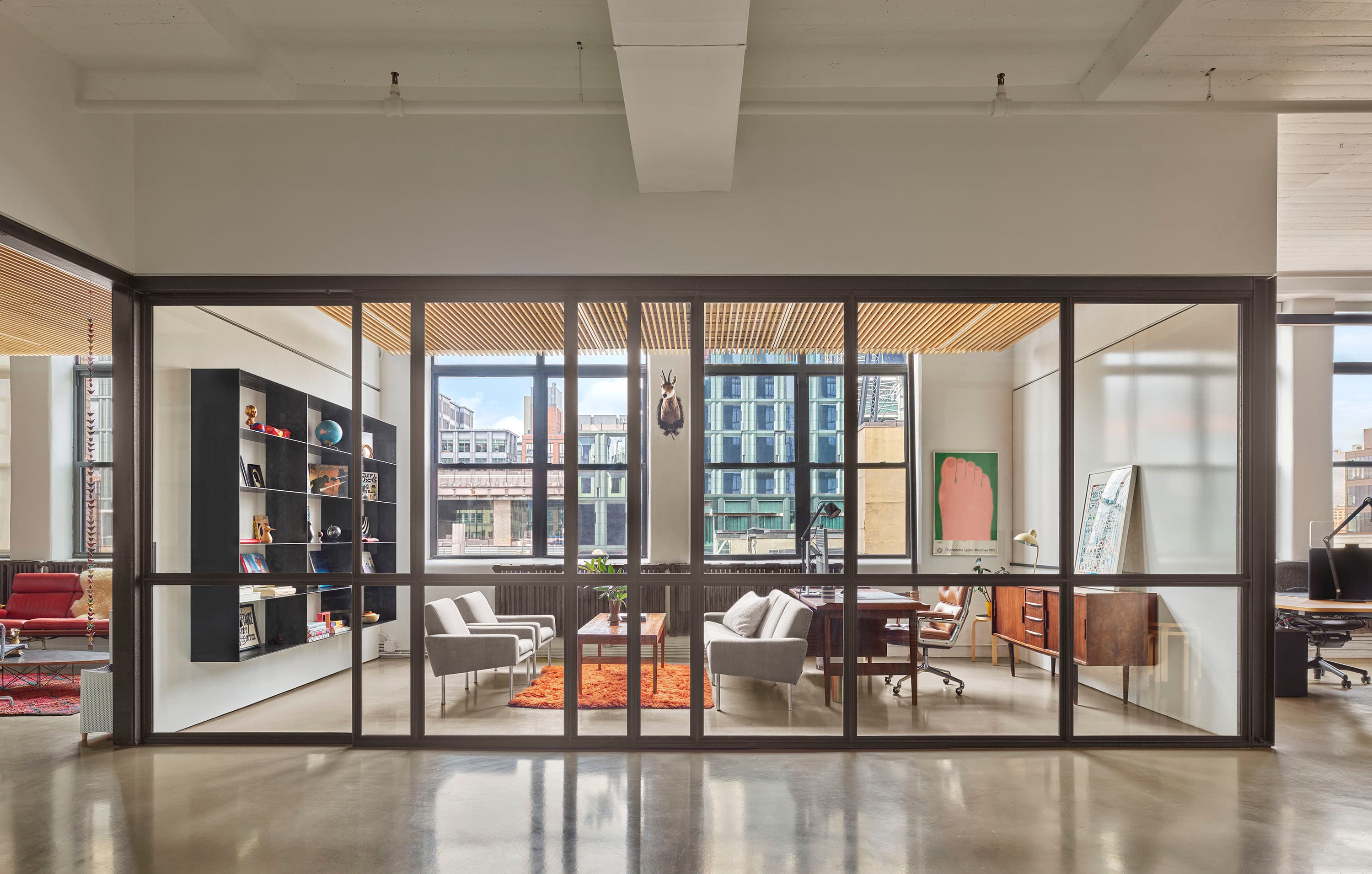 View looking into a glass enclosed office with a bookshelf and two couches and a table in between them on top of an orange rug.